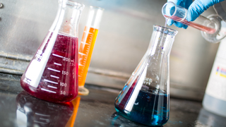 Close up image of a chemistry experiment, with two chemistry flasks, one filled with red liquid and another filled with blue liquid, on a table and a flask filled with an orange liquid in the background. Someone is adding red liquid to the blue liquid flask.