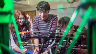 Close up image of a group of students working on a drone. A green light can be seen reflecting off of the glass in front of the students..