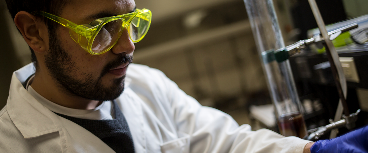 Close up image of a latino male student in a lab coat conducting an experiment.