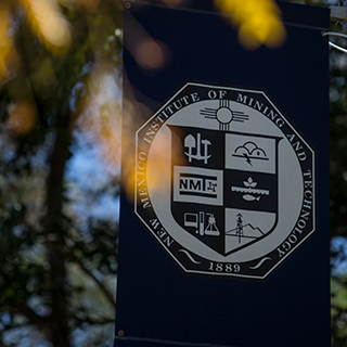 Image of flag with New Mexico Tech Presidential Seal hanging behind a tree.