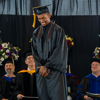 An African-American male dressed in graduation cap and gown looks down and smiles as he prepares to speak during Commencement 2022.