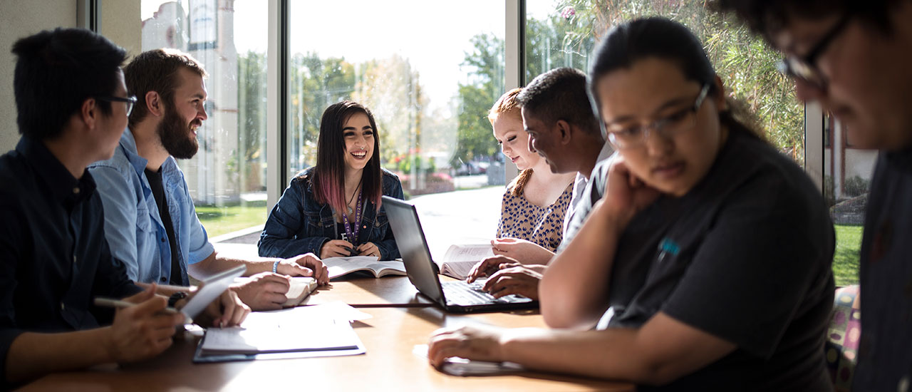 Students sitting at a table studiying together next to large windows. Outside of the windows is a view of greenery and buildings that are present on campus.