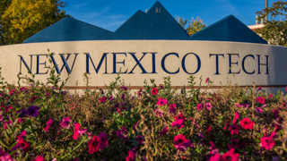 Image of the New Mexico Tech stucco sign entering campus. Red flowers are blooming in the front of the sign.