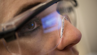 A close up on a researcher's face as she's watching a computer monitor during an experiment. The refection of the monitor can be seen in her safety goggles.