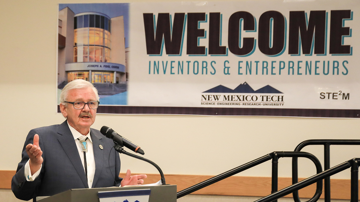Image of NMT President Wells speaking at a podium on the left side of the image. A banner welcoming students to the Inventors and Entrepreneur Workshop is behind him.